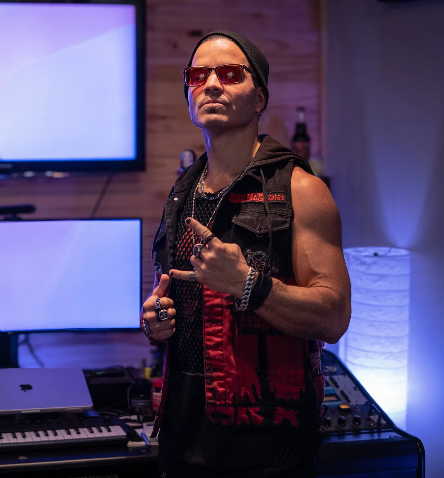 Daemon Grey throws the horns in his home studio, beanie and red-tinted glasses on, wearing a patched battle vest in front of monitors and a keyboard.