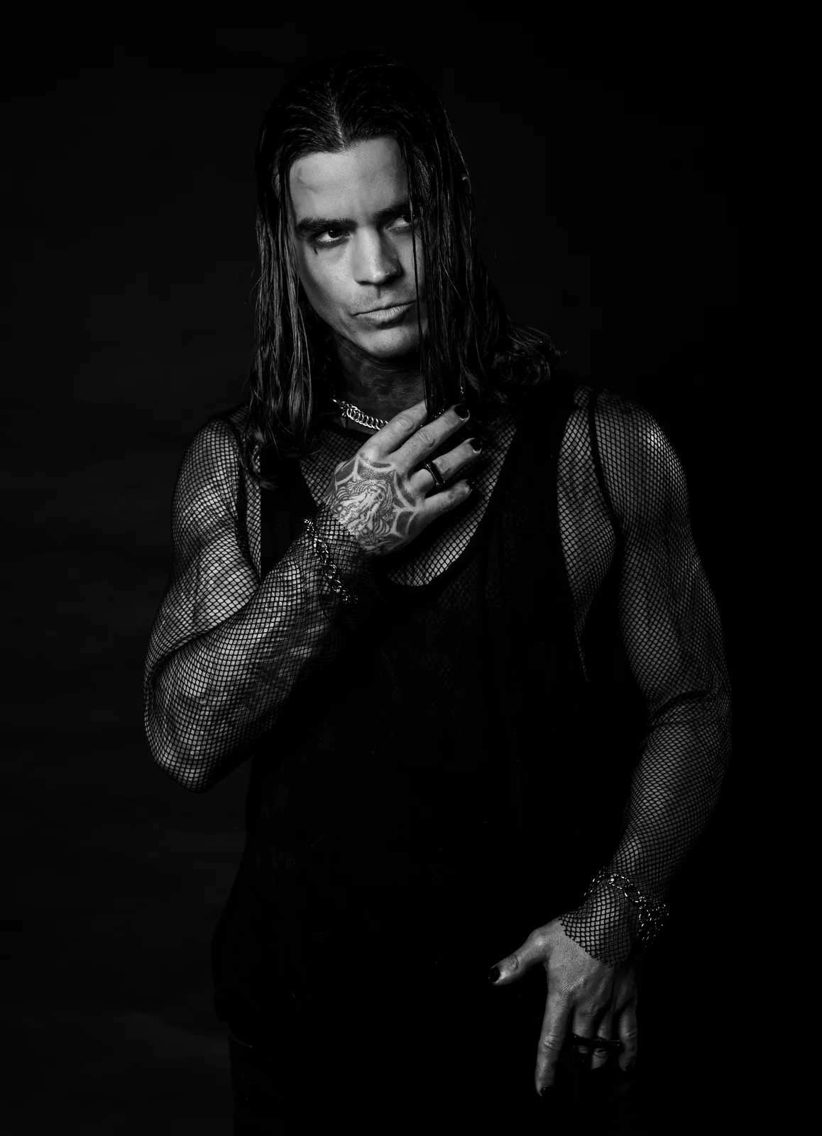 Daemon Grey in a black mesh top, wet hair over one eye, raising a tattooed hand with black nails to his collarbone.
