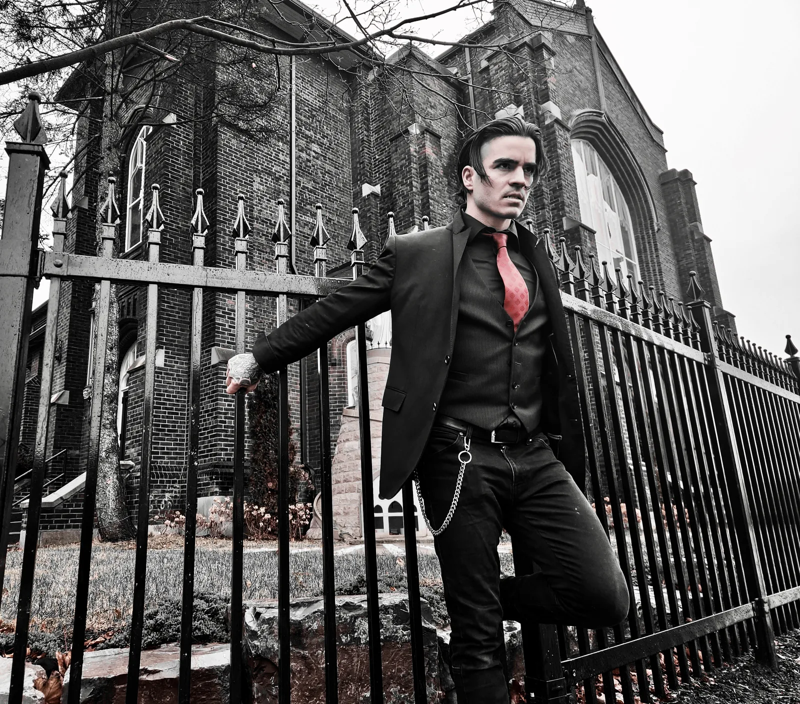Outside a brick church behind a spiked iron fence, everything monochrome except the red tie.