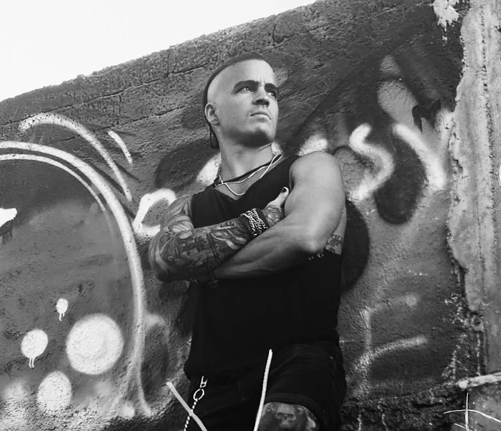 Arms crossed against a graffiti wall in black and white, mohawk sharp, layered chains and a skull tattoo on his shin.