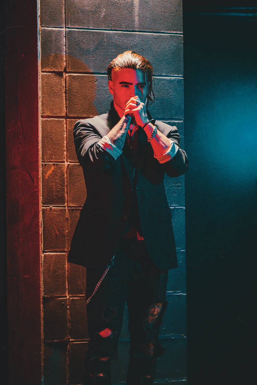 Sharp-suited between a red wall and blue shadow, hair slicked back, hands clasped around the microphone at his lips.