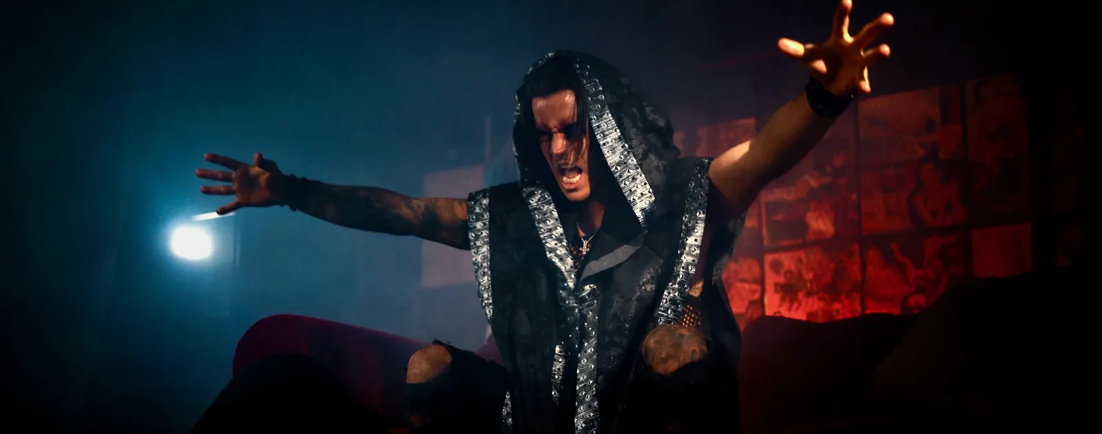 Arms flung wide on the red and blue video set, hooded vest with silver-studded trim, mid-scream over a ruined couch.