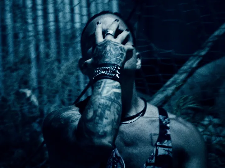 Blue-toned frame of a tattooed, ringed hand clamped over Daemon Grey's face, studded cuff on his wrist.