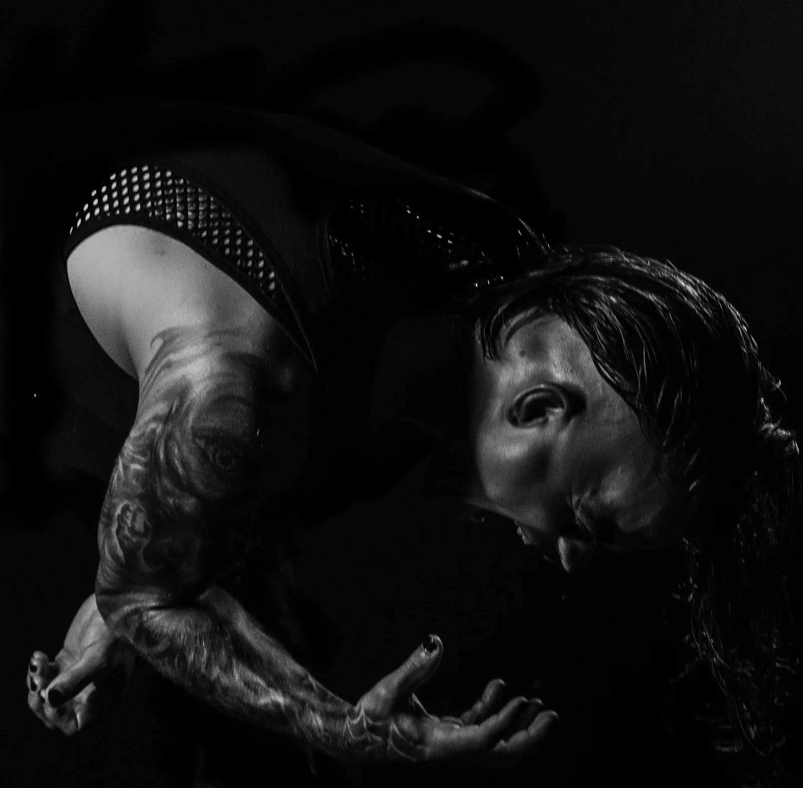 Bent double mid-set in a mesh-panel top, wet hair hanging, hands clawed open under hard side light.