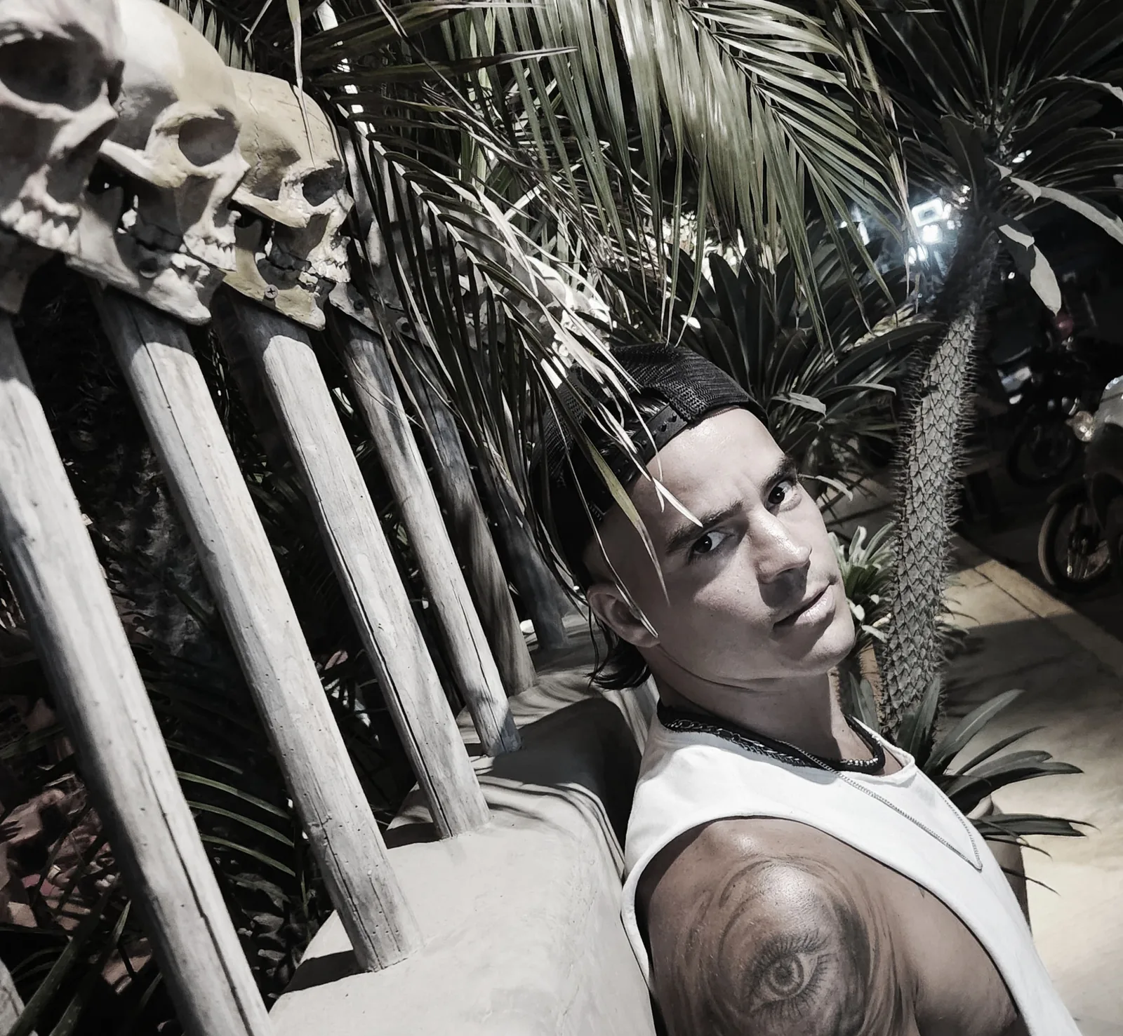 Backwards cap and white tank, Daemon Grey looks over his shoulder from a bench built of carved skulls under palm fronds.