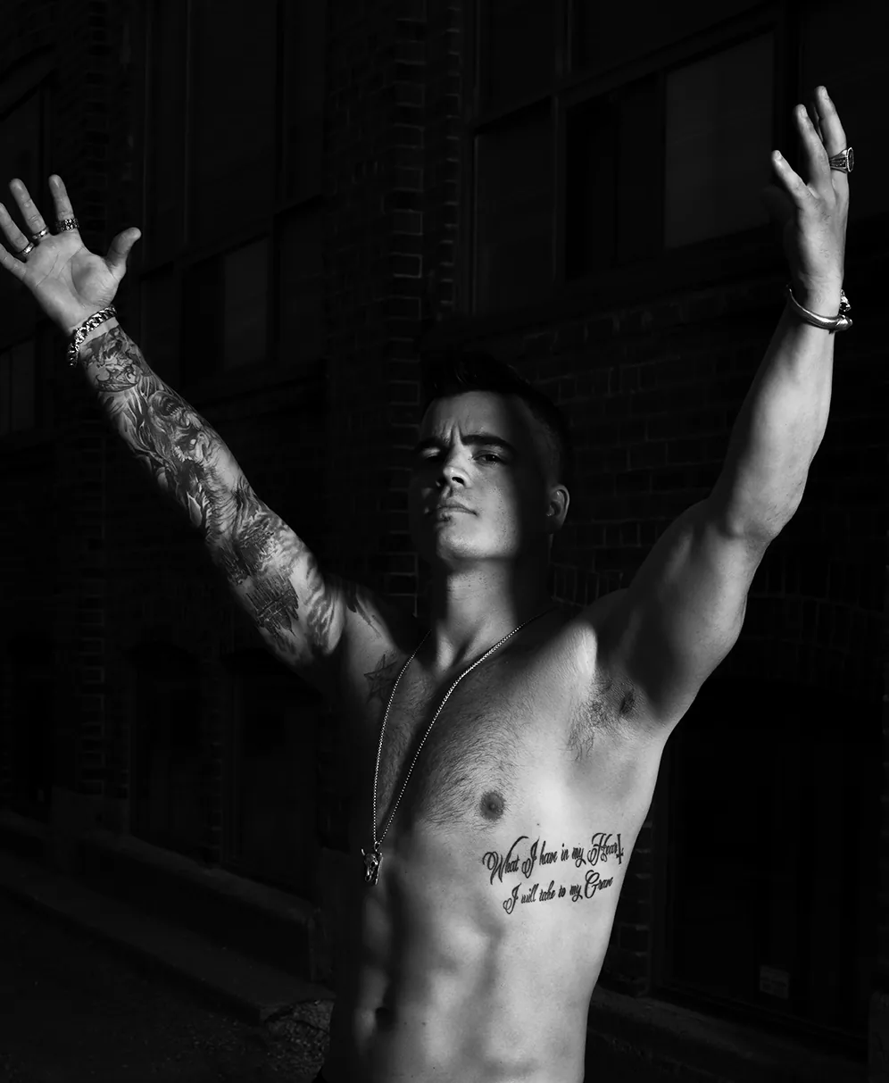 Arms spread wide in a dark alley, shirtless in black and white, the script tattoo across his ribs catching the light.