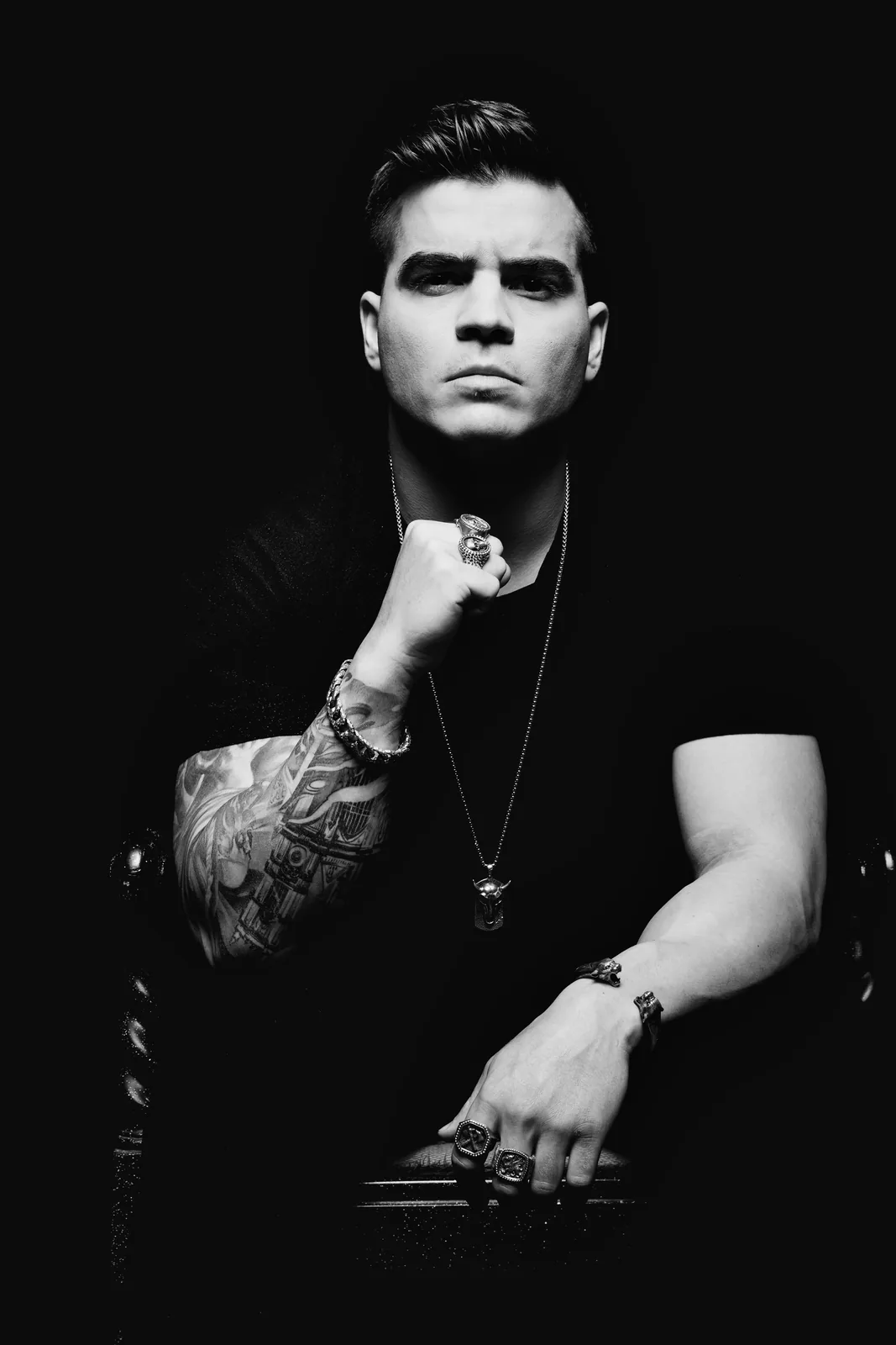 Seated black and white portrait: fist raised under his chin, signet rings and a bull pendant, eyes straight down the lens.