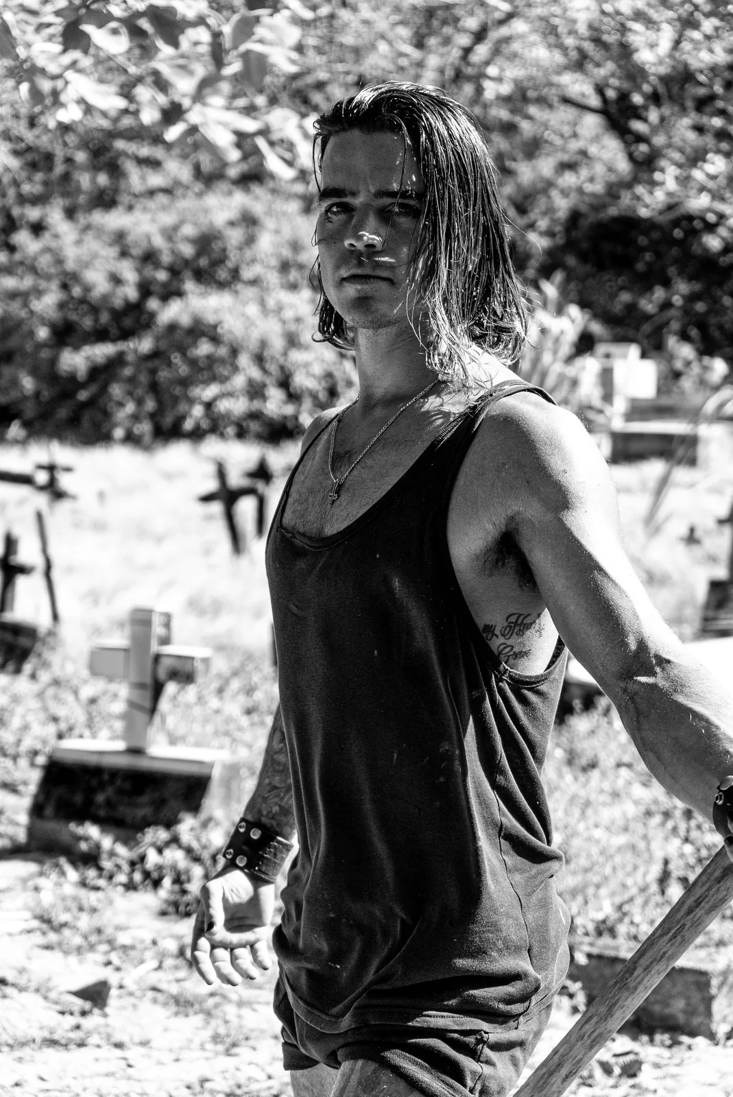 Standing among leaning cross markers in a sunlit cemetery, black tank top and cross pendant, shovel in hand.