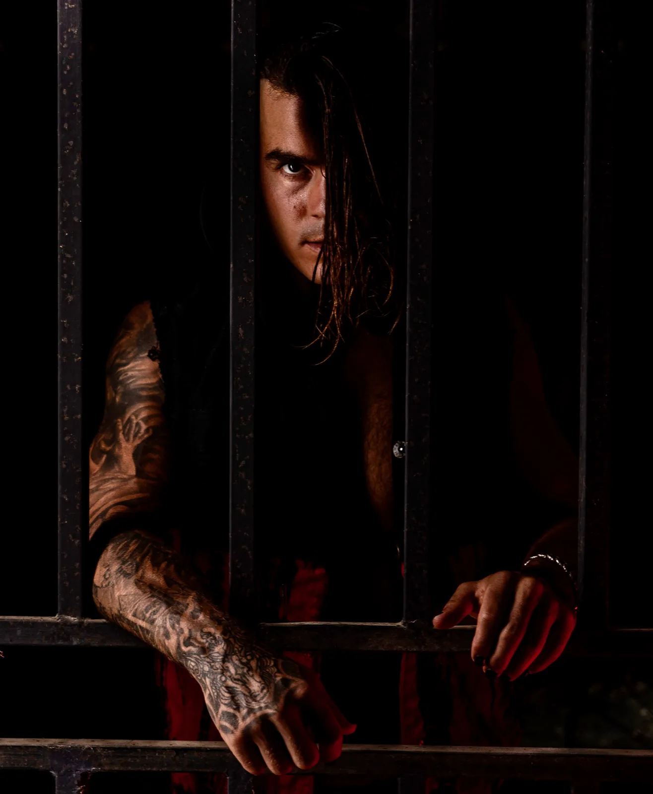 Half in shadow behind a dark iron gate, red light below, one eye fixed on the camera through wet hair.